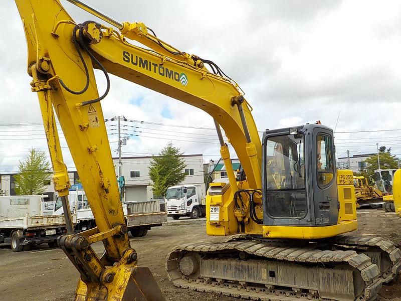 Yellow Sumitomo excavator on a construction site with trucks and buildings in the background.